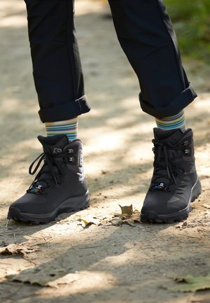 Person trägt schwarze Wanderschuhe und gestreifte Socken, steht auf einem sonnigen Erdweg, umgeben von verstreuten Blättern.