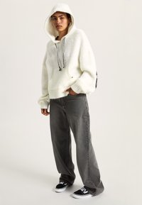 Felpa bianca con cappuccio in maglia spessa, tasca frontale e cordini neri, abbinata a jeans grigi larghi e sneakers nere.