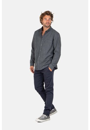 Man standing with one hand in pocket wearing dark gray button-up shirt, navy pants, and gray sneakers on white background.