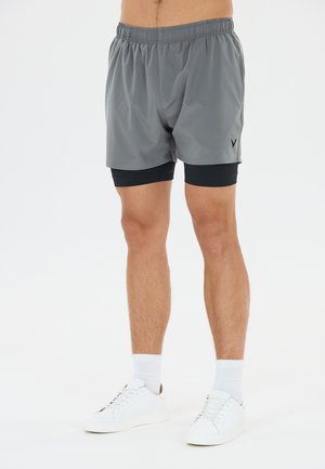 Mand iført grå atletikshorts med sort kompressionslag under, hvide sokker og hvide sneakers, står på en hvid baggrund.