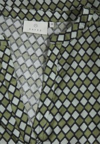 Grønt mønstret stof har et diamantdesign i varierende nuancer, med en V-halskrave og en etiket, der står "KAFFE." Tekstureret overfladedetalje synlig.