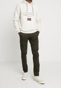 Sudadera gris claro con un logo y un parche de la bandera noruega, combinada con pantalones cargo verdes oliva y zapatillas blancas. Ropa casual para exteriores.