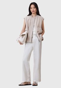 Chaleco sin mangas beige con botones en la parte delantera, camiseta blanca y pantalones de pierna ancha en colores claros. Diseño minimalista, textura suave, combinado con sandalias planas.