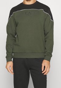 Felpa verde oliva con yoke nero, piping bianco e logo in rilievo sul petto. I polsini e l'orlo a costine conferiscono struttura.