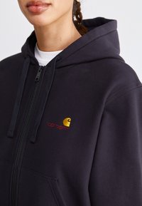 Haut à capuche zippé noir avec une poche kangourou, orné d'un logo brodé doré et rouge sur la poitrine gauche. Fabriqué à partir d'un tissu doux et texturé.