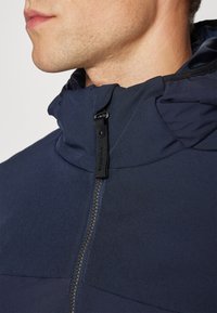 Μπλε ναυτικού σακάκι με ψηλό γιακά, μπροστινό φερμουάρ και αντίθετη υφή υφάσματος στους ώμους, με ρυθμιζόμενο εξοπλισμό.