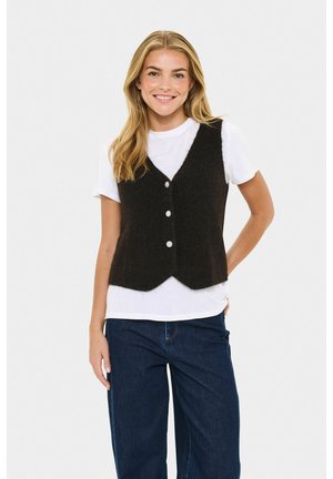 Glunderende vrouw met lang blond haar, gekleed in een wit T-shirt, een zwarte gebreide vest met knopen en donkerblauwe spijkerbroek tegen een effen achtergrond.