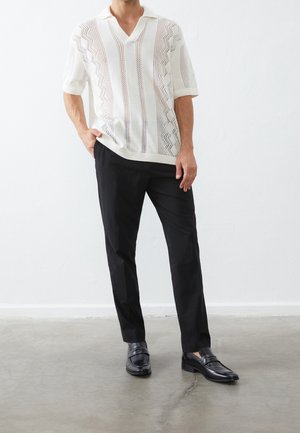 Mužský model oblečený v bílém pleteném tričku s krátkými rukávy a vzory, černých kalhotách na míru a černých kožených mokasínách stojící na betonové podlaze.