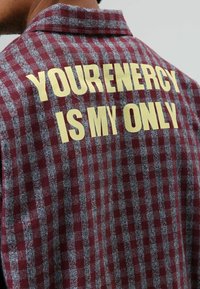 kariertes braunes und graues Stoffhemd mit umgeschlagenem Kragen. Auf der Rückseite befindet sich ein auffälliger gelber Text mit der Aufschrift "YOUR ENERGY IS MY ONLY."