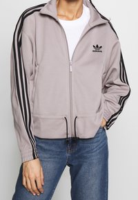 Ljust grå zipjacka i mjukt tyg, med svarta tre-randiga detaljer längs ärmarna och en broderad logotyp på bröstet.