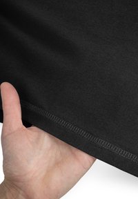 Schwarzer Baumwollstoff mit einer weichen Textur, auf dem eine Hand den Saum hält. Die Nähte sind entlang der unteren Kante sichtbar.