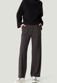 Person trägt dunkelgraue Hosen mit weitem Bein, schwarzen gerippten Rollkragenpullover und silberne metallic Sneaker, steht mit den Händen in den Taschen.