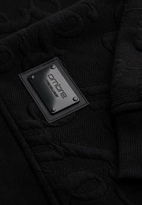 Gros plan sur un tissu noir texturé avec une étiquette métallique rectangulaire noire portant l'inscription « ombre menswear », fixée par quatre rivets.
