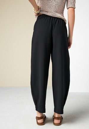 Frau stehend, trägt weite schwarze Hosen, beige Stricktop, Armband aus Holzperlen und braune Sandalen, von hinten gezeigt.