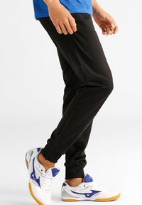 Pantalon de sport noir à coupe slim, doté de poches latérales et de bretelles aux chevilles. Associé à des baskets blanches et bleues, mettant en avant le contraste et le design.