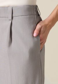 Mano con esmalte de uñas rosa claro descansando dentro del bolsillo de unos pantalones de tiro alto gris claro con cinturilla elástica lateral.