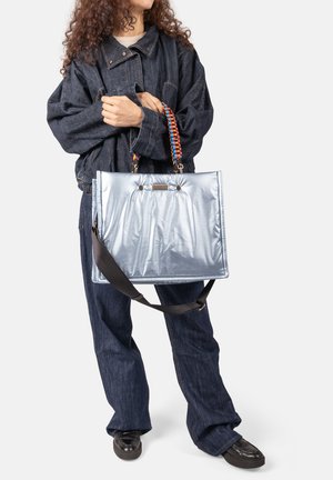 Person in dunklem Denim-Outfit hält große metallic-silberne Tragetasche mit farbig geflochtenem Griff und schwarzem Schulterriemen.