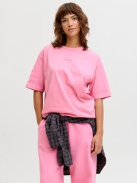Jeune femme aux cheveux bruns ondulés portant un t-shirt rose oversize et un pantalon, avec une chemise à carreaux foncés nouée autour de la taille.