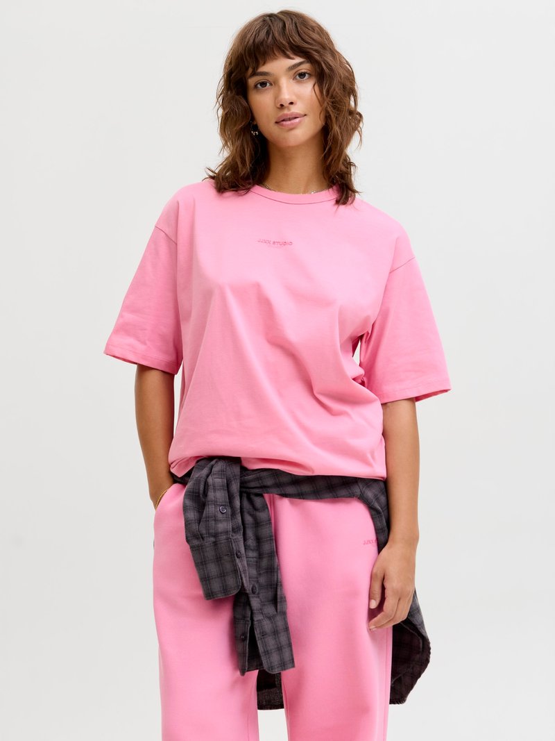 Jeune femme aux cheveux bruns ondulés portant un t-shirt rose oversize et un pantalon, avec une chemise à carreaux foncés nouée autour de la taille.