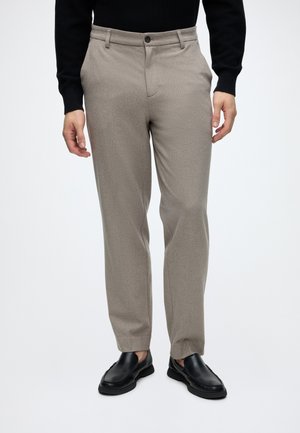 Man wearing light brown tailored trousers with front button and black leather slip-on shoes, standing against a plain background.