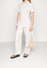 Una persona sta in piedi indossando una camicia a righe leggera a maniche corte e pantaloni larghi, mentre tiene una borsa di perline e indossa sandali neri.