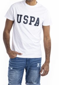 Muž v bílém tričku s nápisem "USPA" a modrých roztrhaných džínových šortkách, stojící s jednou rukou v kapse na bílém pozadí.