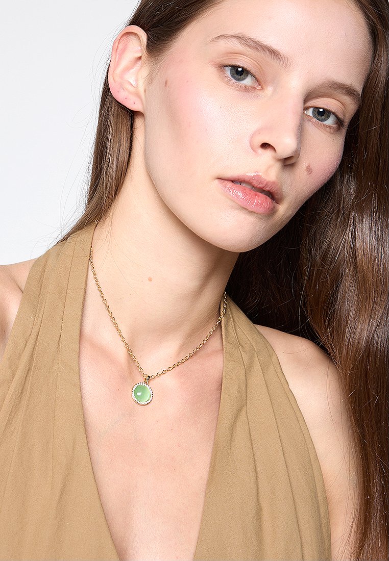 Collar de cadena de oro que presenta un colgante redondo verde en un engaste texturizado. El modelo lleva un top halter beige.