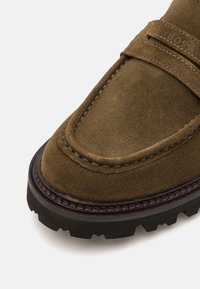 Brown suede slip-on shoe with a thick black rubber sole, stitched edging, and a branded strap across the upper. Textured surface detail.