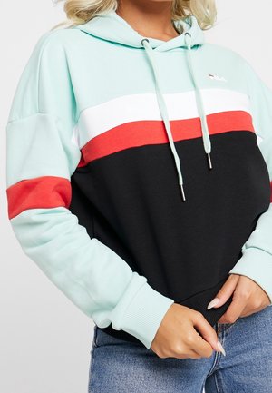 Kobieta nosząca bluzę z kapturem w kolorowe bloki: miętowa zieleń, biel, czerwień i czerń, podciągająca dół bluzy na niebieskie dżinsy.