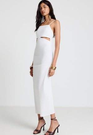 Donna che indossa un vestito midi bianco senza maniche con ritagli laterali, braccialetti dorati e sandali scuri con cinturini e tacco alto, in piedi davanti a uno sfondo semplice.