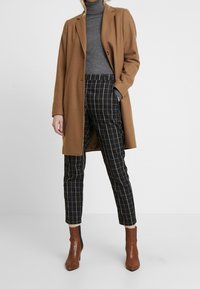 Brown wool-blend coat, gray turtleneck, black and white checkered trousers, and brown ankle boots with a pointed toe. Smooth texture and tailored fit.
