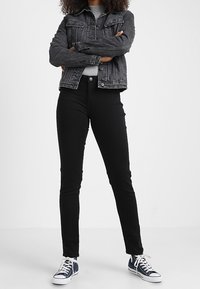 Černé slim džíny spojené se šedou džínovou bundou s předními kapsami a zapínáním na knoflíky, nošené s modrými teniskami.