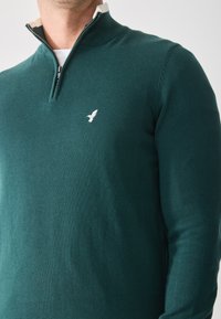 Pulover tricotat verde cu guler cu jumătate de fermoar, având un accent alb sub guler și un mic logo alb al unui păun pe piept.
