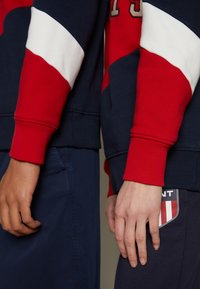 Due felpe con colori a contrasto: una a righe rosse e bianche su fondo blu navy, entrambe con polsini a coste. Un'etichetta con logo è visibile sul capo inferiore.