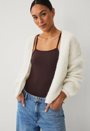 Frau mit dunklen Haaren in einer weißen, flauschigen Strickjacke, braunem ärmellosem Top und blauen Jeans, die mit den Händen in den Taschen vor einem schlichten Hintergrund steht.