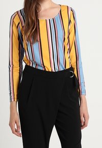 Långärmad skjorta med flerfärgade vertikala ränder i gult, blått och rosa, tillsammans med högmidjade svarta byxor som har en rund bältesspänne.