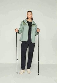 Naine kannab rohelist jope, musti pükse ja beeže kingi, hoides käes kahte musta matkakeppi, seistes ühtlasel heledal taustal.