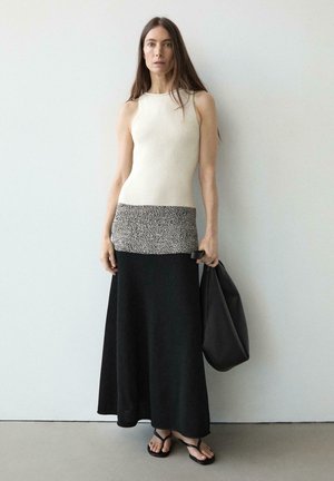 Donna con lunghi capelli castani che indossa un top beige senza maniche, gonna bianca e nera, sandali neri, tiene una grande borsa nera, in piedi contro un muro semplice.