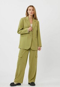 Blazer entallado en verde oliva con un solo botón y dos bolsillos frontales, combinado con pantalones cargo a juego que tienen bolsillos laterales. Zapatos negros.