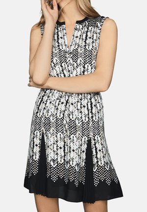 Kobieta nosząca czarno-białą sukienkę bez rękawów z geometrycznym wzorem, plisowanymi panelami i dekoltem w serek.