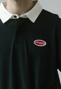 Dark green polo shirt with a white collar, featuring a red embroidered logo on an oval patch. Soft texture and classic design.
