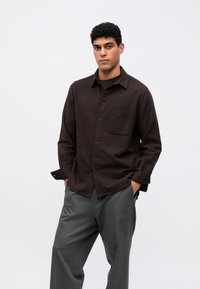 Camicia marrone con bottoni, maniche lunghe e una tasca sul petto, abbinata a pantaloni verdi a gamba larga, posizionata su uno sfondo semplice.