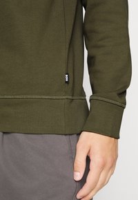 Sudadera verde oliva con puños y dobladillo acanalados, con una etiqueta negra de "BOSS" en el lateral, fabricada en un material suave y texturizado.