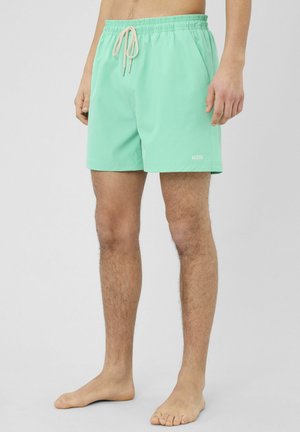 Homme portant un short de bain vert menthe avec un cordon blanc et un petit logo blanc "BZB" sur la cuisse gauche, debout pieds nus devant un fond uni.