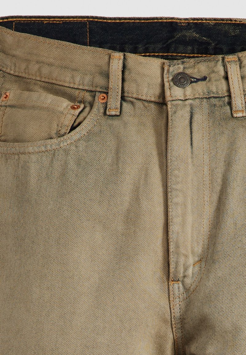 Jeans de mezclilla beige con cierre de botón, remaches de cobre y costuras de contraste. Presenta un diseño tradicional de cinco bolsillos y un ligero desgaste.