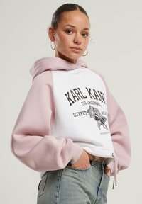 Biely skrátený hoodie s ružovými rukávmi, čierny grafický potlačový text a dizajn orla na prednej strane, rebrované manžety a nastaviteľný šnúrkový detail.