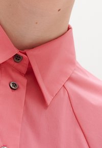 Camicia col colletto rosa chiaro con design a bottoni, texture liscia e bottoni scuri a contrasto. Vestibilità su misura con colletto strutturato.