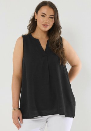 Donna con capelli castani lunghi che indossa una canottiera nera senza maniche e pantaloni bianchi, in piedi contro uno sfondo chiaro e uniforme.