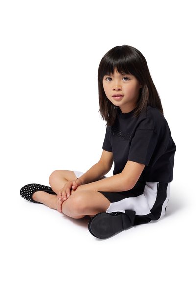 T-shirt cropped noir avec détail de logo, shorts noir et blanc, et chaussures noires à enfiler avec des accents perforés. Enfant assis les jambes croisées.