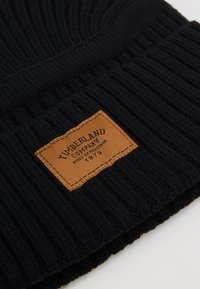 Musta ribattu kudottu pipot, jossa on ruskea nahkaleima, jossa lukee "Timberland Company" ja "Built on Heritage 1973" painettuna.